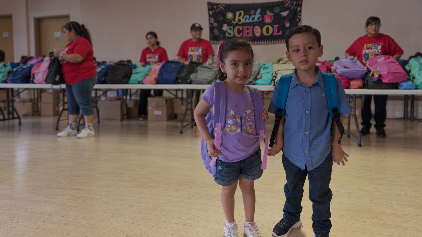 Two students at a back-to-school backpack giveaway with colorful backpacks and volunteers in the background