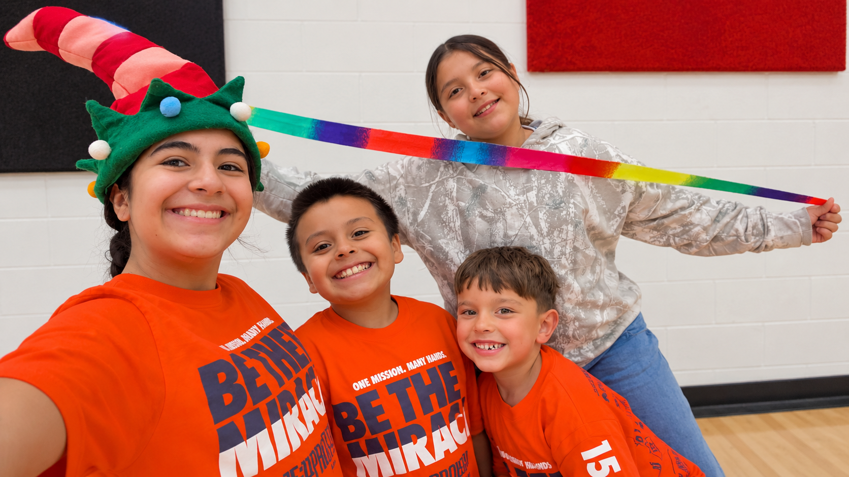 Three children smiling together indoors while holding a rainbow ribbon during a student activity
